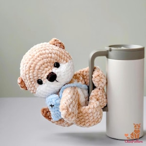 May include: A handmade, plush otter toy in shades of tan and white, with a blue bib, clinging to the handle of a white travel mug. The otter has black eyes and a brown nose. The mug has a grey handle and lid.