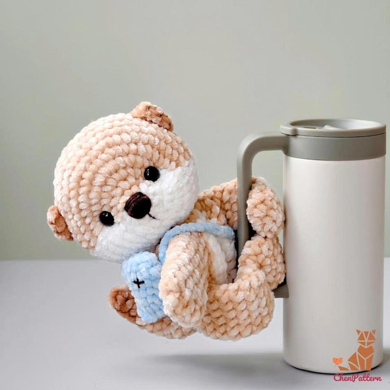 May include: A handmade, plush otter toy in shades of tan and white, with a blue bib, clinging to the handle of a white travel mug. The otter has black eyes and a brown nose. The mug has a grey handle and lid.