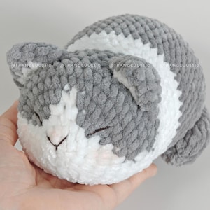 May include: A plush, crocheted cat toy featuring a soft, textured yarn in alternating patches of grey and white. The cat has closed eyes, a small triangular nose, and rounded ears. The text @TRANGLUU2310 is visible across the top of the toy.