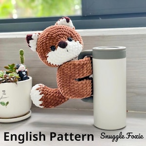 Può includere: Una volpe marrone all'uncinetto con dettagli bianchi, che tiene in mano un bicchiere da viaggio bianco e grigio. La volpe ha occhi neri e un muso bianco. Il testo "English Pattern Snuggle Foxie" è in basso.