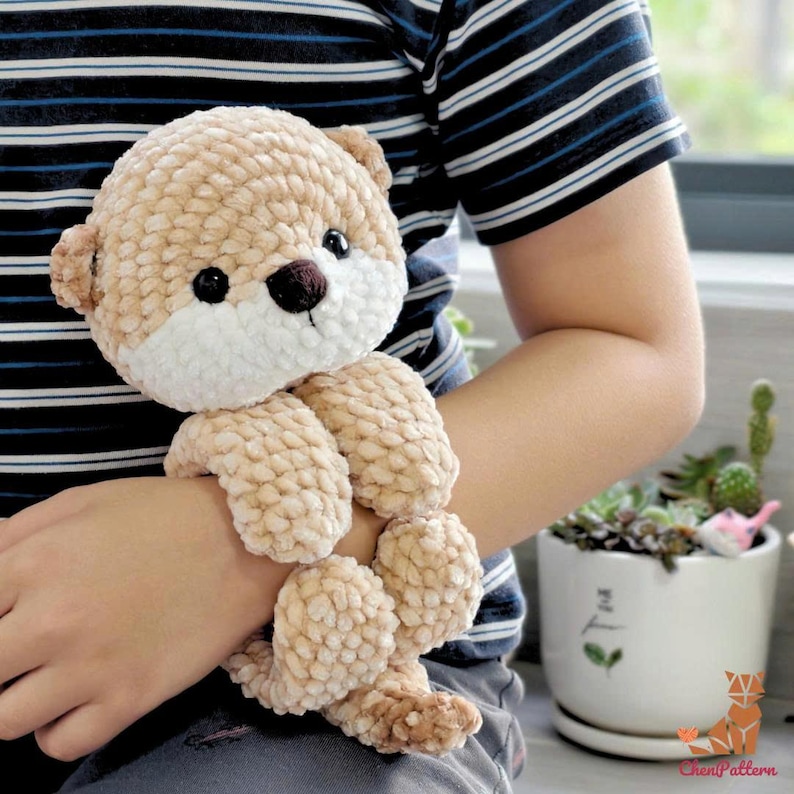 May include: A crocheted teddy bear in beige and cream tones. The bear features a white face, dark eyes, and a brown nose. Its arms and legs are designed to wrap around a wrist. The bear is made from soft, textured yarn, perfect as a cuddly toy.