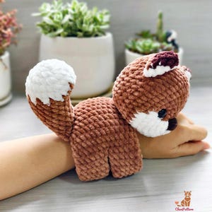 May include: A crocheted fox toy in shades of brown and white. The fox has a fluffy white tail and a small, dark nose. The fox is lying on its belly. The toy is handmade and has a soft, textured appearance.