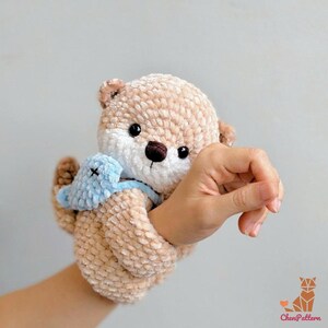 May include: A crocheted otter toy in light brown with a white face and a small blue toy. The otter has black eyes and a brown nose. The toy is held by a hand.
