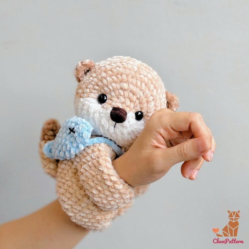 May include: A crocheted otter toy in light brown with a white face and a small blue toy. The otter has black eyes and a brown nose. The toy is held by a hand.