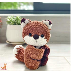 May include: A handmade crochet fox plushie in shades of brown, white, and burgundy. The fox has a fluffy texture, with a white belly, face, and tail tip. It has small black eyes and a dark brown nose. The fox is sitting down.