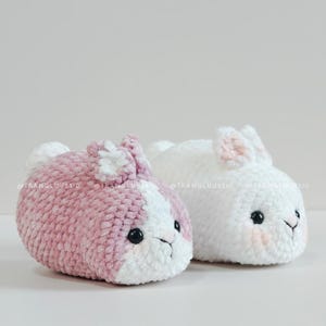 May include: Two plush crochet bunnies, one in dusty pink and white and the other in solid white. Both feature black plastic eyes, small stitched noses, and upright ears. The text @TRANGLUU2310 is visible across the center of the image.