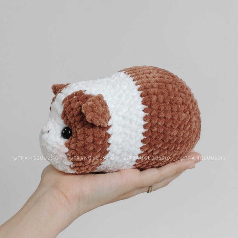 May include: A small, plush, crocheted guinea pig toy featuring a soft, textured yarn in alternating bands of brown and white. The stuffed animal has two small, dark, round eyes and tiny, rounded ears, with the text @TRANGLUU2310 visible.