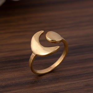 Könnte beinhalten: Ein goldfarbener Ring mit einem Halbmond-Design und einem kleinen, runden Akzent. Der Ring besteht aus einem glänzenden, metallischen Material und ist auf einer dunklen Holzoberfläche platziert. Das Design ist schlicht und elegant.