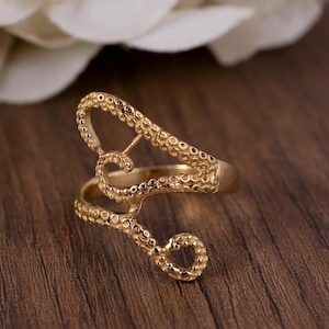 Könnte beinhalten: Ein goldfarbener Ring mit einem Oktopus-Tentakel-Design. Der Ring hat ein wirbelndes, offenes Design mit detaillierten Saugnapfstrukturen. Der Ring befindet sich vor einem verschwommenen Hintergrund aus einer Holzoberfläche und einer weißen Blume.