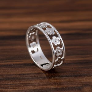 Könnte beinhalten: Ein silberner Ring mit floralem Design. Der Ring hat ein Band mit ausgeschnittenen Blumenformen. Der Ring besteht aus glänzendem Metall und wird auf einer dunkelbraunen Holzoberfläche präsentiert.