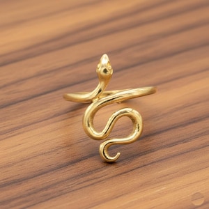 Könnte beinhalten: Ein goldfarbener Ring in Form einer Schlange. Der Kopf der Schlange ist erhoben, und ihr Körper windet sich um den Finger. Der Ring besteht aus einem glänzenden, metallischen Material und ist vor einem Holzgrund platziert.
