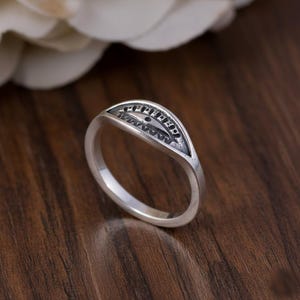 Könnte beinhalten: Ein silberner Ring mit einem bogenförmigen Design. Der Ring hat ein detailliertes Muster innerhalb des Bogens, einschließlich Text und kleiner geometrischer Formen. Der Ring liegt auf einer dunklen Holzoberfläche.