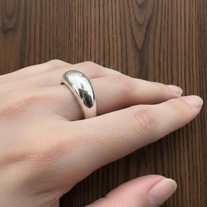 Könnte beinhalten: Ein polierter, silberner Ring mit einem glatten, abgerundeten Design. Der Ring wird an einem Finger getragen und reflektiert das Licht. Der schlichte, elegante Stil des Rings macht ihn alltagstauglich.