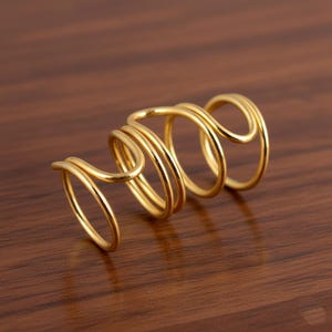 Könnte beinhalten: Ein goldfarbener Ring mit einem einzigartigen Spiraldesign. Der Ring hat mehrere Schlaufen und Kurven, die eine auffällige und moderne Ästhetik erzeugen. Das Schmuckstück befindet sich vor einem warmen Holzuntergrund.
