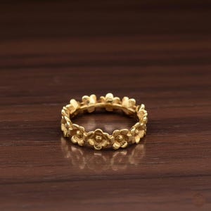 May include: A gold-colored ring featuring a continuous floral design. The ring is made of a shiny, metallic material and is set against a dark brown wooden surface. The flowers are small and detailed, creating a delicate and elegant look.