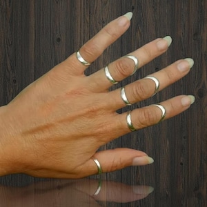 May include: A hand wearing several silver rings. The rings are of various designs, including simple bands and open-ended styles that curve around the fingers. The background is a dark wood grain.