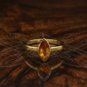 Könnte beinhalten: Ein goldener Ring mit einem marquiseförmigen, orange-gelben Edelstein. Der Ring hat ein schlichtes, poliertes Goldband. Der Edelstein hat einen Farbverlauf von tiefem Orange zu hellem Gelb. Der Ring wird auf einer dunklen, gemusterten Oberfläche präsentiert.