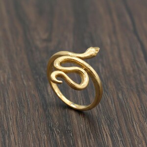 Könnte beinhalten: Ein goldfarbener Ring in Form einer Schlange. Die Schlange windet sich um den Finger, mit erhobenem Kopf. Der Ring besteht aus einem glänzenden, metallischen Material und liegt auf einer dunklen Holzoberfläche.
