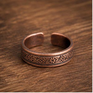 Könnte beinhalten: Ein kupferfarbener, offener Ring mit einem dunklen, vertieften Blumen- und Rankenmuster, das in die Mitte eingraviert ist. Die Kanten des Rings sind mit einem feinen, sich wiederholenden Schraffurmuster versehen, das dem Metall ein gealtertes Aussehen verleiht.