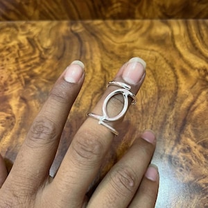 May include: A silver-colored ring with an oval-shaped design and two bands. The ring is worn on a finger, and the background is a wooden surface.