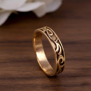 Könnte beinhalten: Ein goldfarbener Metallring mit einem durchgehenden, erhabenen, rankenartigen Wirbelmuster, das sich um das äußere Band windet. Die vertieften Bereiche zwischen den erhabenen goldfarbenen Metalldesigns sind dunkel und erzeugen einen Kontrast.