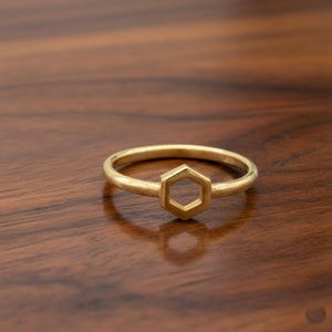 May include: A gold-colored ring with a hexagonal design. The ring is set against a dark brown wooden surface. The ring's band is thin, and the hexagonal shape is the focal point of the ring.