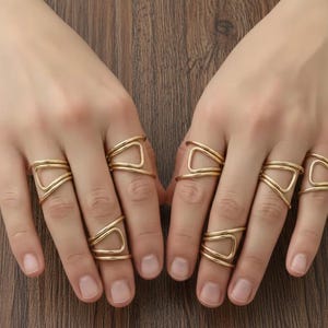 May include: Gold-colored rings with a geometric design are displayed on two hands. Each ring features a unique open shape, creating a modern and stylish look. The rings are made of a shiny, metallic material.