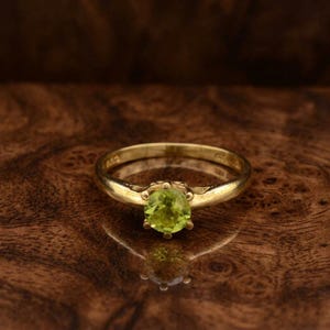 Könnte beinhalten: Ein goldener Ring mit einem runden, hellgrünen Edelstein. Der Ring hat ein schlichtes Band und eine Vier-Krappen-Fassung. Der Edelstein ist der Mittelpunkt, vor einem warmen, hölzernen Hintergrund.