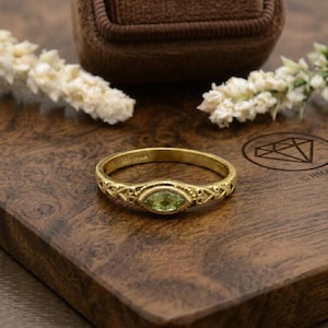 Könnte beinhalten: Ein goldener Ring mit einem ovalen, hellgrünen Edelstein. Der Ring hat ein aufwendiges, durchbrochenes Design am Band. Der Ring wird auf einer Holzoberfläche mit einer braunen Ringschatulle im Hintergrund präsentiert.