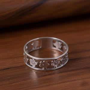 Könnte beinhalten: Ein silberner Ring mit floralem Design. Der Ring hat eine Reihe von ausgeschnittenen Blumenformen um das Band. Der Ring besteht aus einem glänzenden, metallischen Material und ist vor einem warmen, braunen Hintergrund platziert.