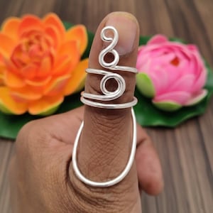 May include: A silver-colored thumb ring with a unique, open design. The ring features a spiral and circular detail on the top of the thumb, with a curved band extending down the side. The background includes orange and pink flowers.