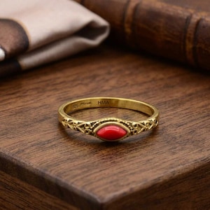 Könnte beinhalten: Ein goldfarbener Ring mit einem ovalen, roten Edelstein in der Mitte. Der Ring hat ein verziertes Filigran-Design und die Aufschrift "925" und "HMA". Der Ring liegt auf einer dunklen Holzoberfläche.
