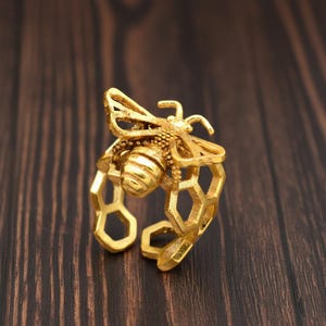 Könnte beinhalten: Ein goldfarbener Ring mit einem detaillierten Bienen-Design. Die Biene sitzt auf einem bandförmigen Wabenmuster. Der Ring ist aus Metall gefertigt und hat eine strukturierte Oberfläche, die die komplizierten Details des Insekts zeigt.