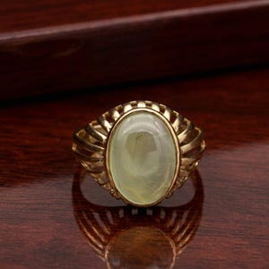 Könnte beinhalten: Ein goldfarbener Ring mit einem ovalen, hellgrünen Edelstein. Der Ring hat ein detailliertes, strukturiertes Band mit einem geriffelten Design um den Edelstein. Der Edelstein ist etwa 2 cm lang.