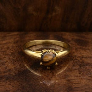 Könnte beinhalten: Ein goldfarbener Ring mit einem runden, braunen und goldfarbenen Edelstein. Der Ring hat ein glattes, poliertes Band, und der Edelstein ist in einer sechszackigen Fassung gefasst. Der Ring wird auf einer dunklen Holzoberfläche präsentiert.