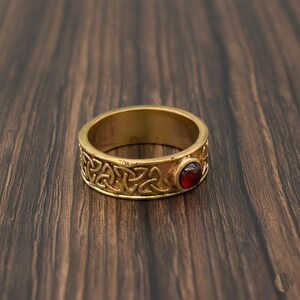 Könnte beinhalten: Ein goldfarbener Ring mit keltischem Knotenmuster und einem roten Edelstein. Der Ring ist vor einem dunklen Holzmaserungshintergrund platziert, der die filigranen Details des Metalls und die leuchtende Farbe des Steins hervorhebt.