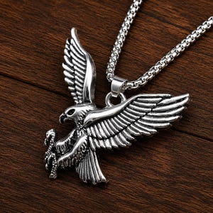 Puede incluir: Un colgante de metal plateado con forma de águila en vuelo, que presenta plumas detalladas en las alas y garras que sujetan un pequeño objeto. El colgante está unido a un collar de cadena tipo caja de color plateado con acabado pulido.