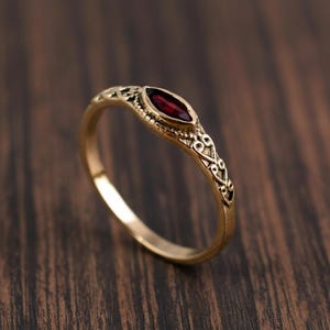 Könnte beinhalten: Ein goldfarbener Ring mit einem ovalen, tiefroten Edelstein. Das Band weist aufwendige, dekorative Muster auf. Der Ring ist vor einem dunklen Holzmaserungshintergrund platziert, der die Details hervorhebt.