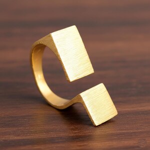 Könnte beinhalten: Ein goldfarbener Ring mit modernem, geometrischem Design. Der Ring hat zwei rechteckige Elemente, die durch ein gebogenes Band verbunden sind. Der Ring besteht aus gebürstetem Metall und liegt auf einer Holzoberfläche.