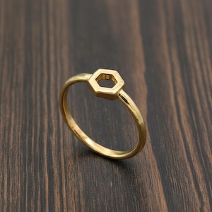 Könnte beinhalten: Ein goldfarbener Ring mit sechseckigem Design in der Mitte. Der Ring befindet sich vor einem dunklen Holzmaserungshintergrund. Das Ringband ist dünn und glatt, und das sechseckige Mittelstück hat ein ausgehöhltes Design.