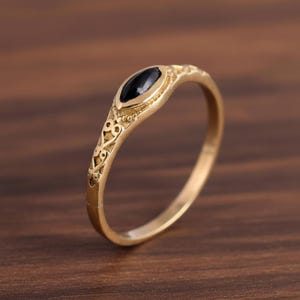 Könnte beinhalten: Ein goldfarbener Ring mit einem ovalen, schwarzen Edelstein. Der Ring hat ein verziertes, graviertes Design am Band. Der Ring liegt auf einer dunkelbraunen Holzoberfläche.