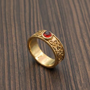 Könnte beinhalten: Ein goldfarbener Ring mit einem roten Edelstein in der Mitte und einem keltischen Knotenmuster auf dem Band. Der Ring liegt auf einer dunklen Holzoberfläche. Das Design ist detailliert.