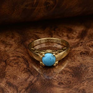 Könnte beinhalten: Goldfarbener Ring mit einem runden, hellblauen Stein in der Mitte. Der Ring hat ein verziertes Design am Band und der Text "GTQ-1002" ist sichtbar. Der Ring ist vor einem dunklen Hintergrund mit Holzmaserung platziert.