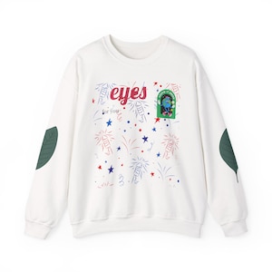 Puede incluir: Sudadera blanca con un diseño gráfico que presenta la palabra "eyes" en rojo, fuegos artificiales, estrellas y un personaje de dibujos animados. Las mangas tienen parches de hojas verdes.