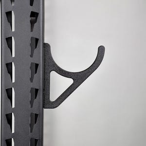 May include: A matte black metal J-hook attachment secured to a vertical rack upright. The upright features a series of evenly spaced, trapezoidal cutouts along its length, while the J-hook provides a curved cradle for supporting gym equipment.
