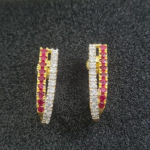 Op de afbeelding: Een paar goudkleurige oorringen. Elke oorring heeft een rij kleine, ronde, rode edelstenen en een rij heldere, witte edelstenen. De oorbellen zijn geplaatst tegen een donkergrijze achtergrond.