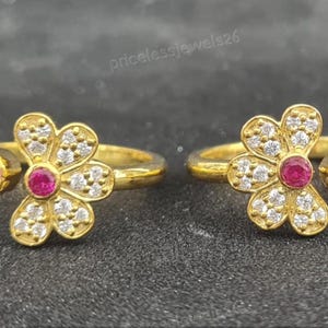 May include: Two gold toe rings. Each ring has a flower design with a central red gemstone and surrounding white gemstones. A smaller red gemstone is set on the band. The rings are displayed on a dark surface.