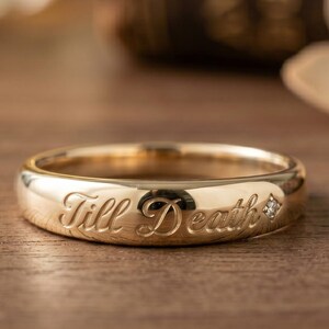 May include: A gold ring with the words "Till Death" engraved around the band. A small diamond is set into the ring. The ring is displayed on a wooden surface.