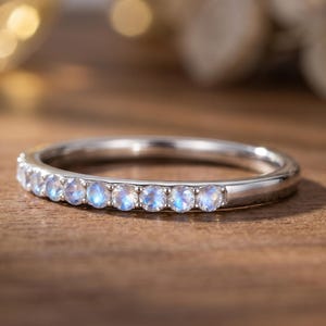 May include: A silver ring featuring a line of small, round, pale blue gemstones. The ring sits on a wooden surface, with a soft, blurred background. The gemstones are uniformly spaced and reflect light.
