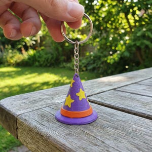 May include: A purple wizard hat keychain with yellow star accents and an orange band. The keychain is attached to a silver chain and ring. The hat is sitting on a wooden table.